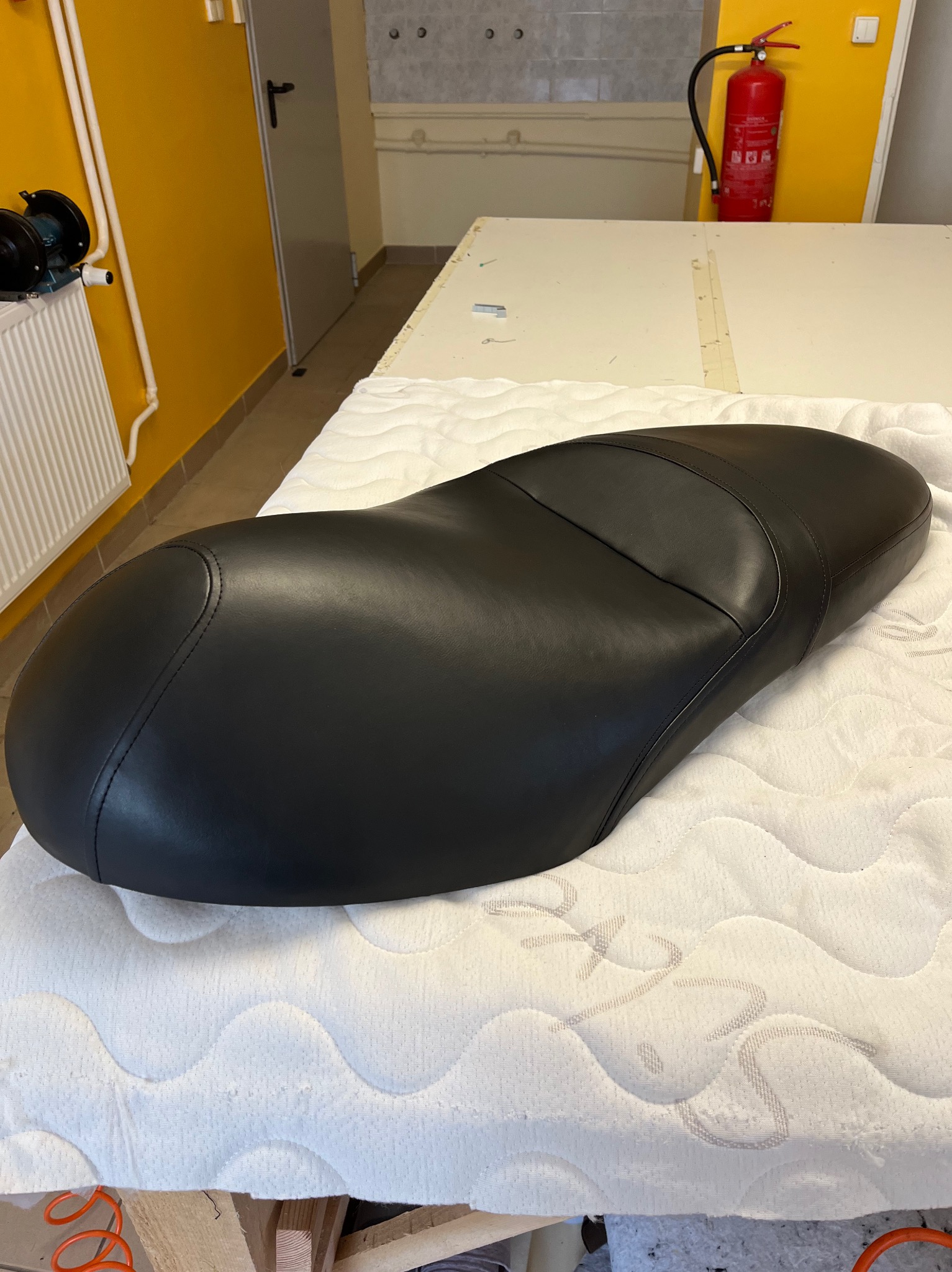 Czarne skórzane siedzisko motocyklowe leży na białej pikowanej powierzchni, prawdopodobnie podczas procesu naprawy lub odnawiania.