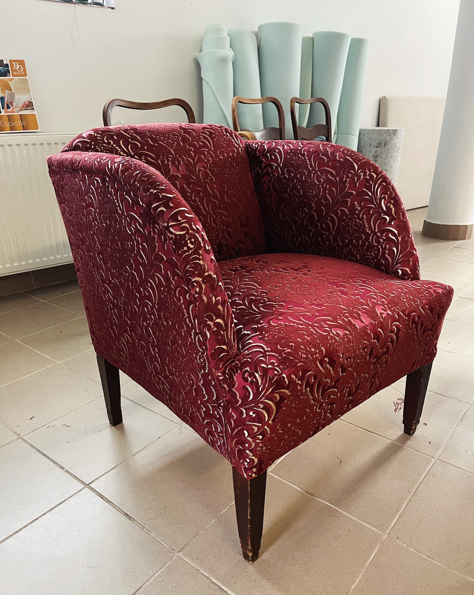 Odnowiony fotel w bordowej tkaninie z delikatnym, złotym wzorem, stojący na kafelkowej podłodze, w tle widoczne rolki z pianką tapicerską i szkielety innych mebli.