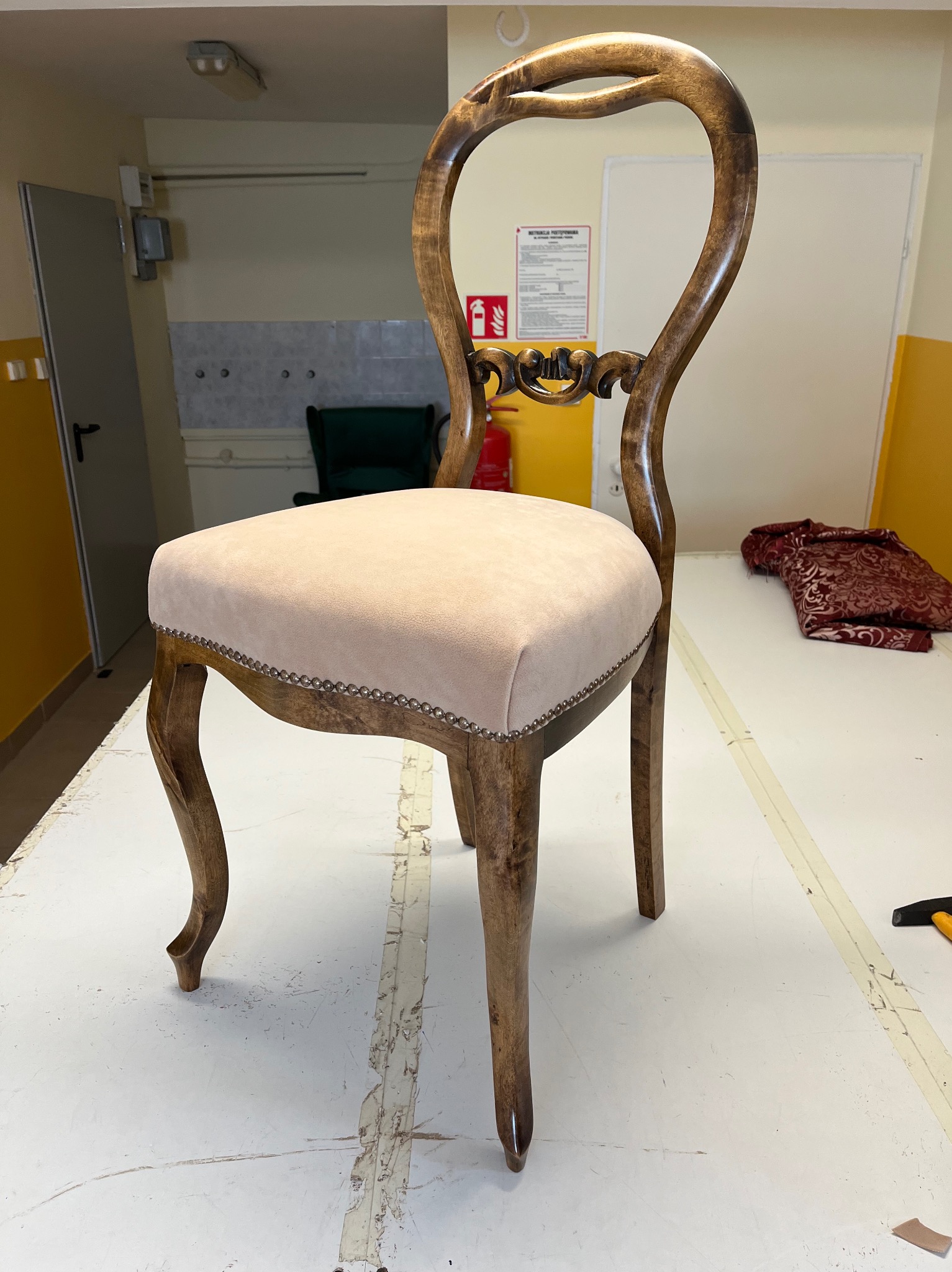 Krzesło po renowacji tapicerskiej z nowym, jasnoróżowym siedziskiem zdobionym srebrnymi pineskami, stojące w warsztacie tapicerskim na stole roboczym.