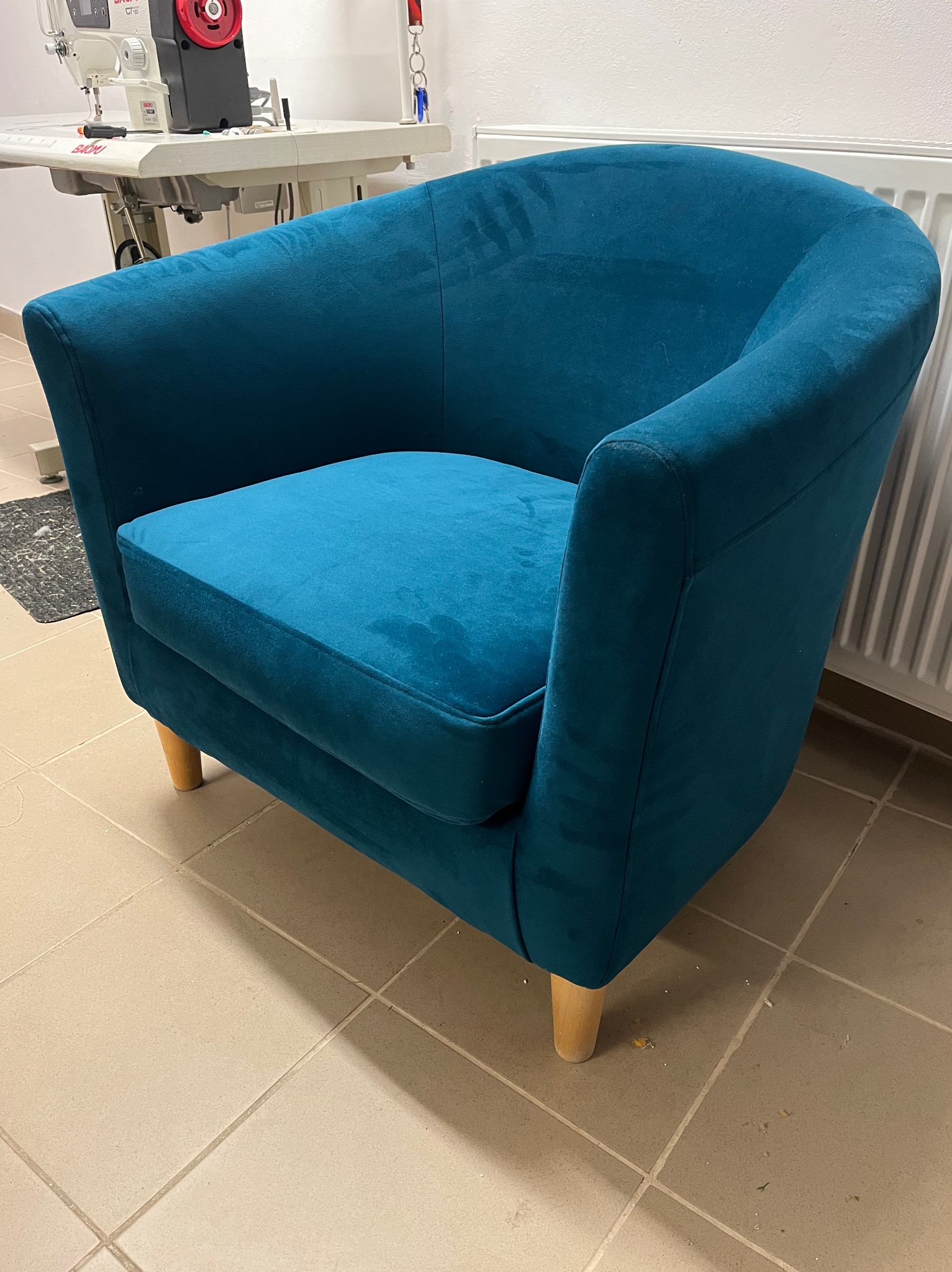 Turkusowy fotel klubowy z drewnianymi nóżkami, stojący na kafelkach, z widoczną maszyną do szycia w tle.