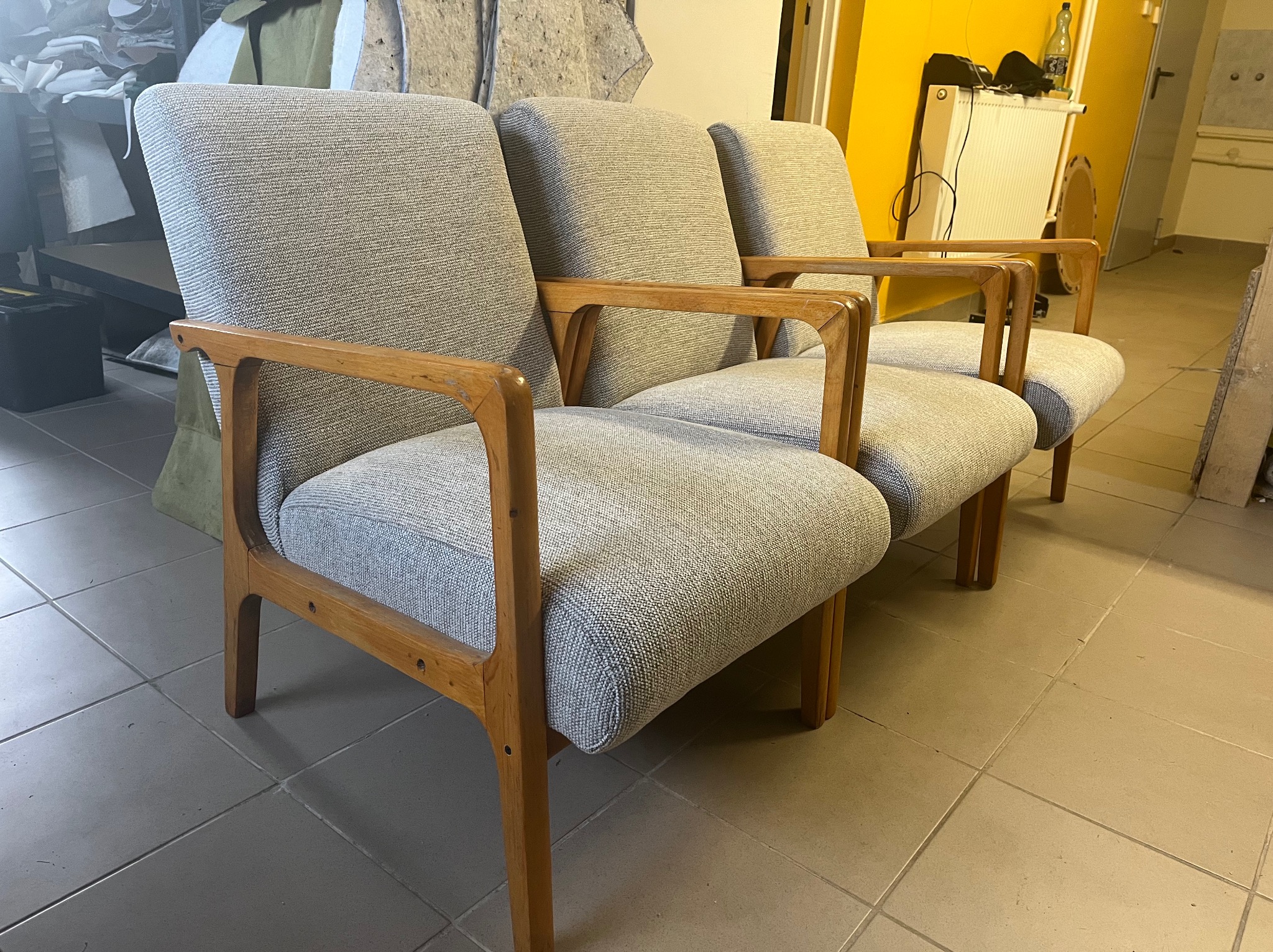 Trzy odnowione fotele w stylu vintage z drewnianymi ramami i szarą tapicerką, ustawione jeden za drugim na kafelkowej podłodze w warsztacie.