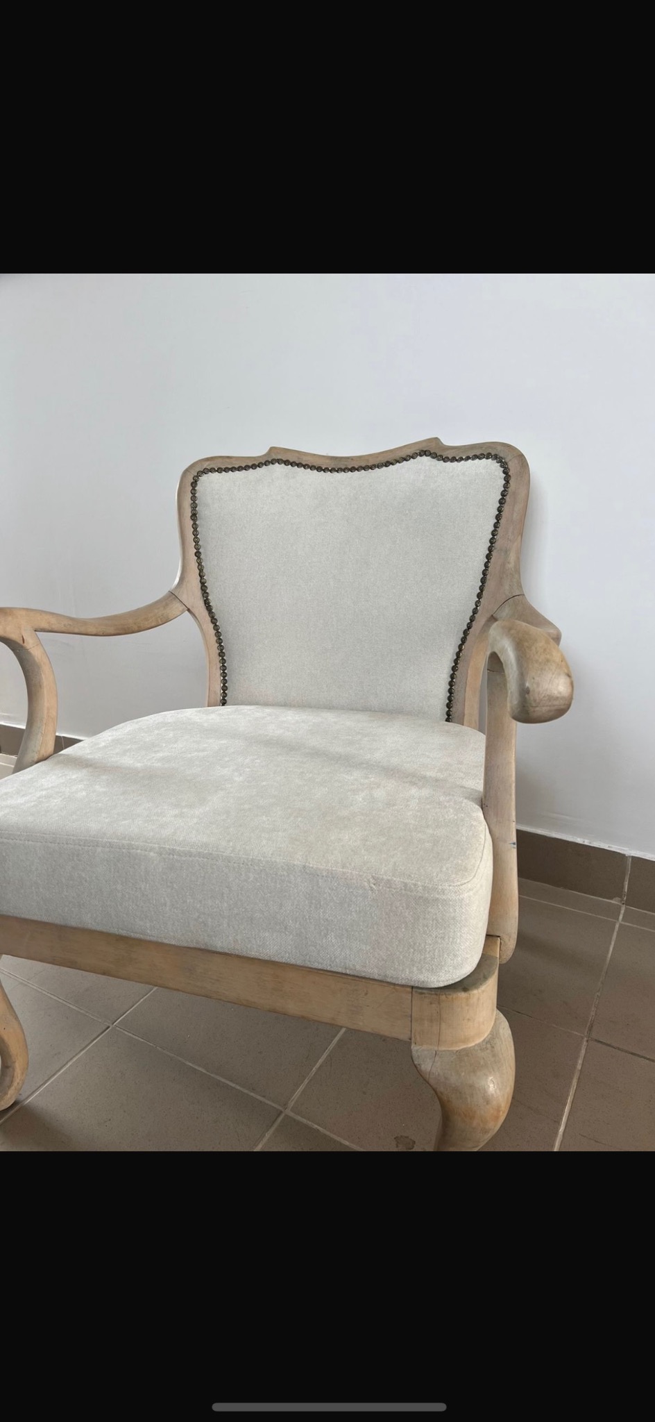 Odrestaurowany fotel z jasnym drewnem i kremową tapicerką, wykończony ozdobnymi ćwiekami, stojący na brązowej podłodze z płytek.