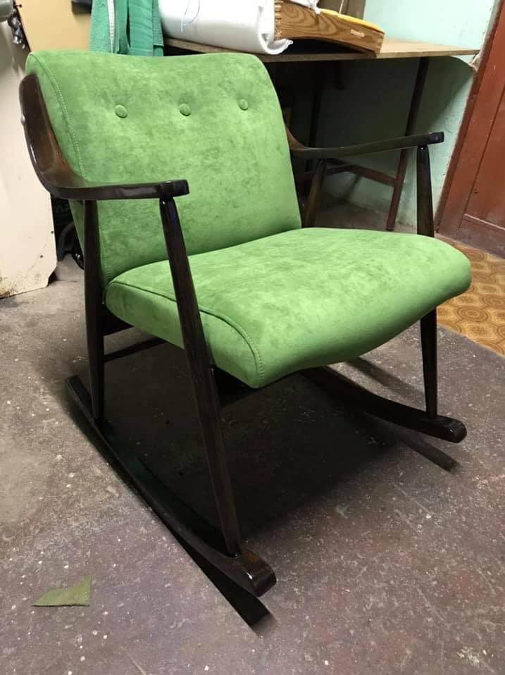 Odnowiony fotel bujany z ciemnego drewna i jasnozieloną tapicerką, widoczne trzy guziki na oparciu i fragment odciętej tapicerki na podłodze.