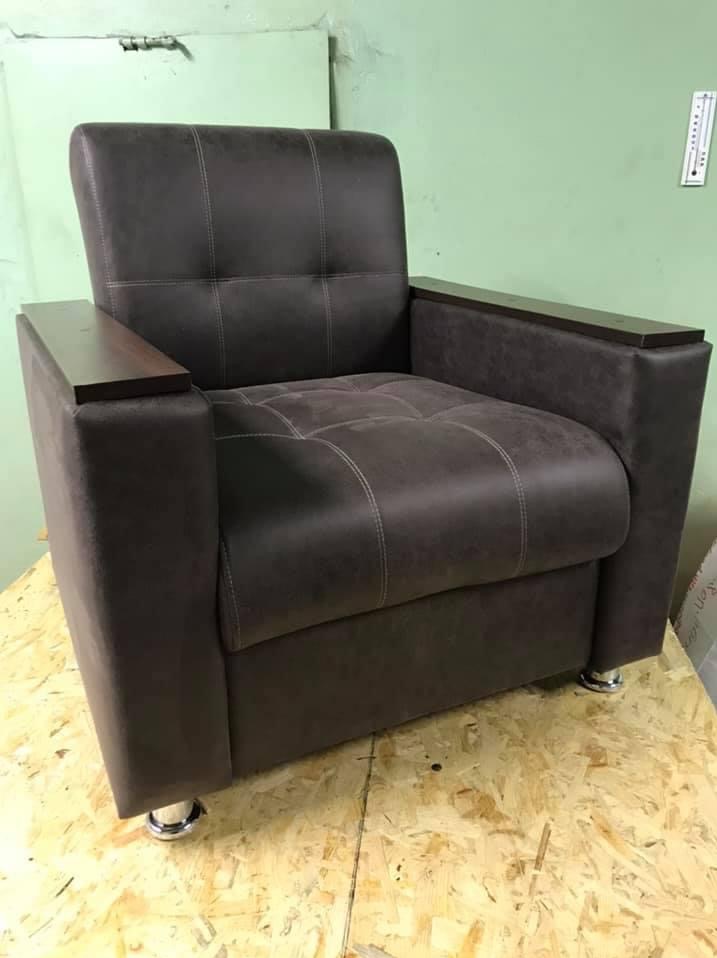 Brązowy fotel z drewnianymi podłokietnikami i chromowanymi nóżkami, widoczny na tle zielonej ściany i płyty OSB.