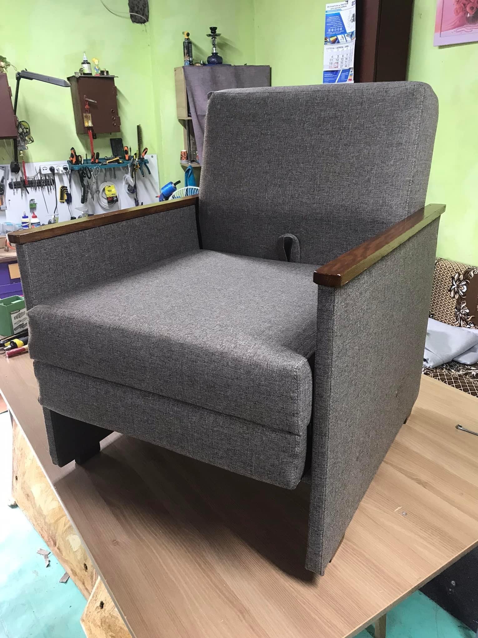 Szary, tapicerowany fotel z drewnianymi podłokietnikami, stojący na stole w warsztacie z narzędziami na ścianie.