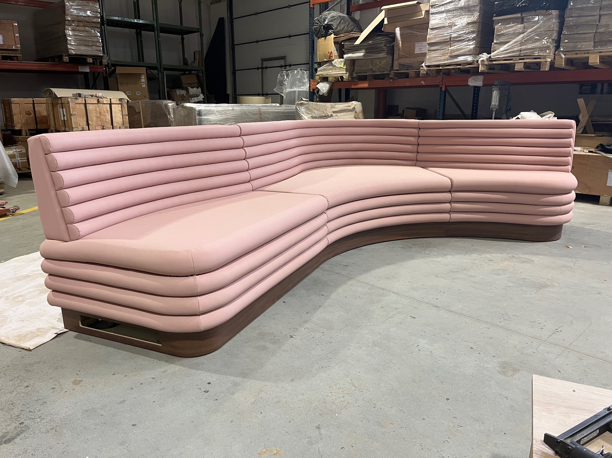 Narożnik tapicerowany na różowo z poziomymi przeszyciami, na drewnianej podstawie, sfotografowany w magazynie z regałami wypełnionymi kartonami w tle.