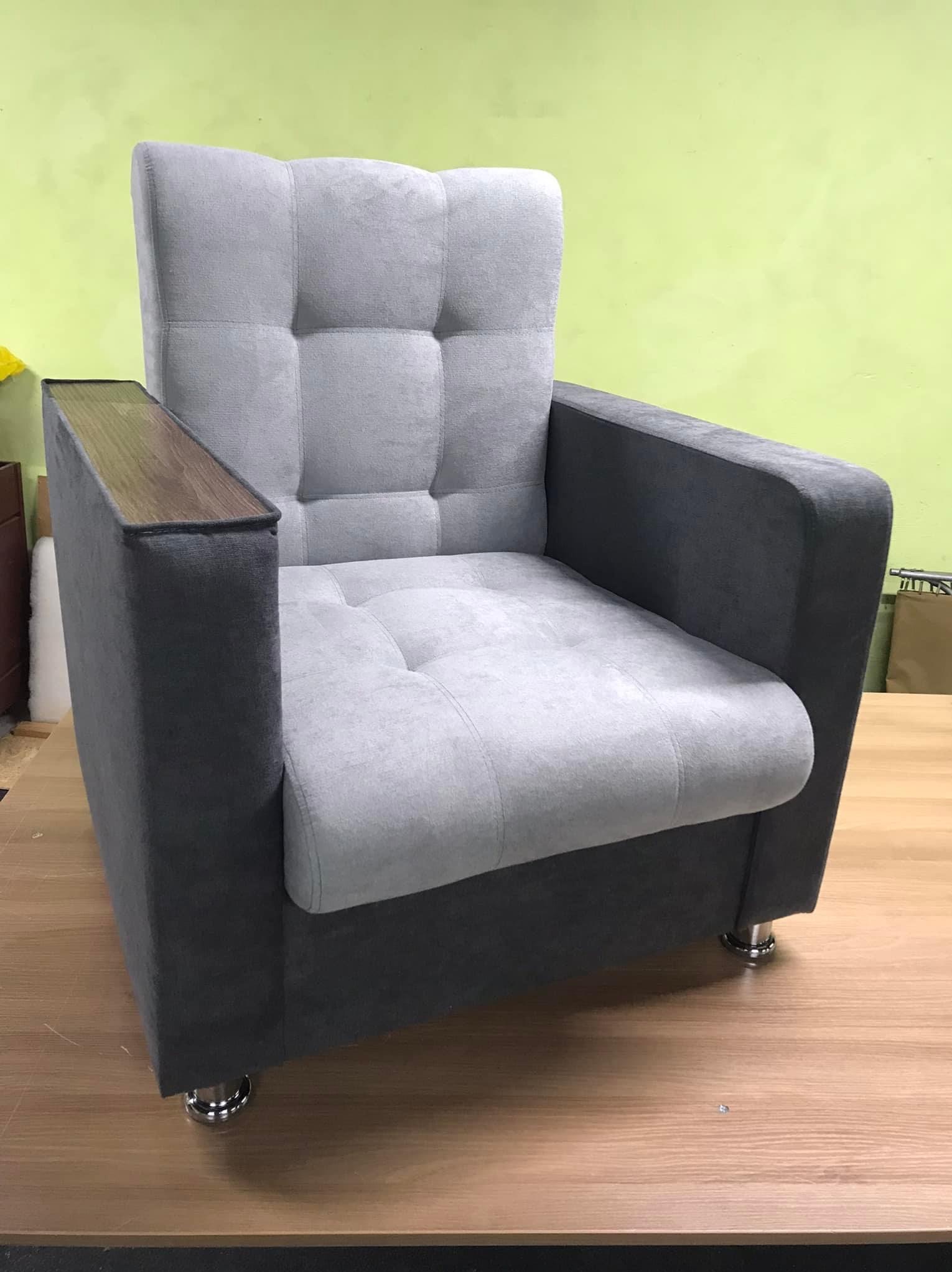 Szary fotel tapicerowany z drewnianymi podłokietnikami i pikowanymi poduszkami na siedzisku i oparciu, stojący na drewnianym blacie na tle zielonej ściany.