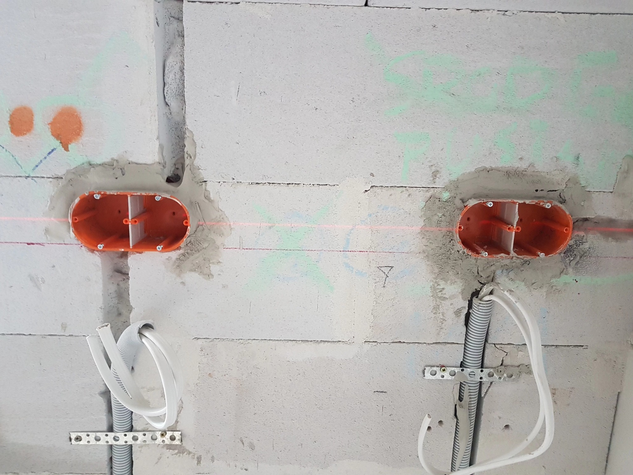 Dwa pomarańczowe gniazda elektryczne podtynkowe zamontowane w szarym bloku betonowym, z widocznymi przewodami w peszlu i poziomą linią lasera wyznaczającą poziom.