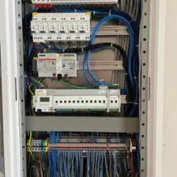 Rozdzielnica elektryczna z licznymi przewodami, zabezpieczeniami i modułami, z opisami kabli, widok z bliska na instalację w szafie.