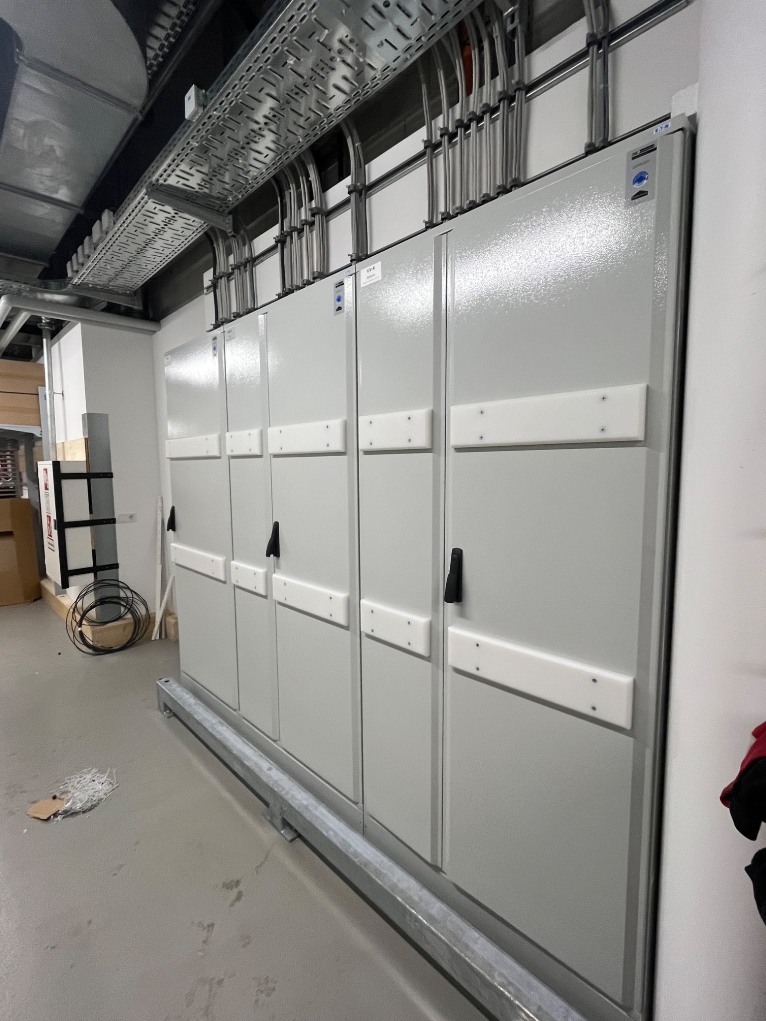 Szara rozdzielnia elektryczna z rzędami przewodów w metalowych korytkach pod sufitem, widok z dołu, kable w peszlach.