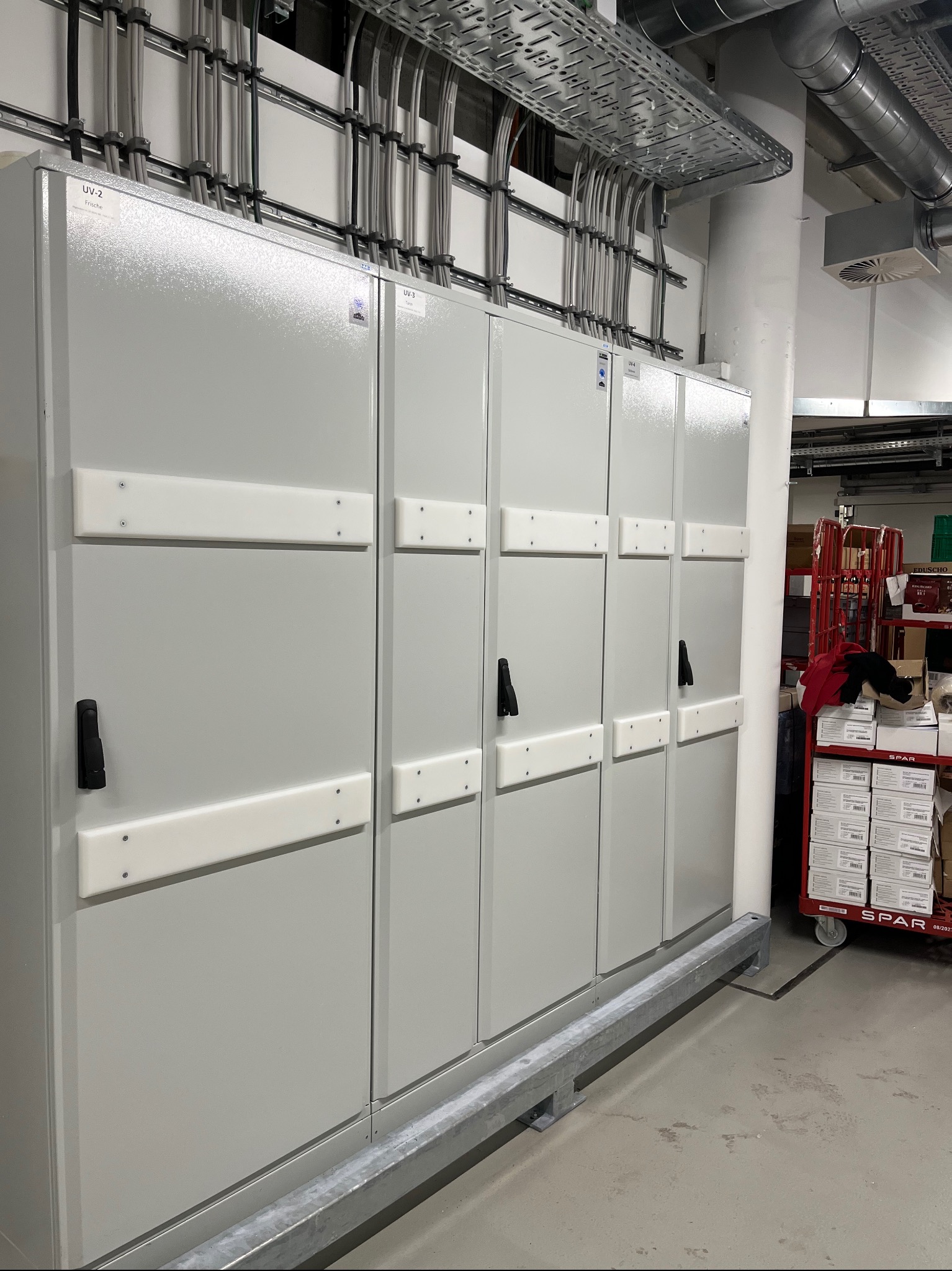 Szereg szaf elektrycznych UV-2 do UV-6 w szarej obudowie, z widocznymi kablami w korytkach nad szafami, umieszczonych w pomieszczeniu technicznym.