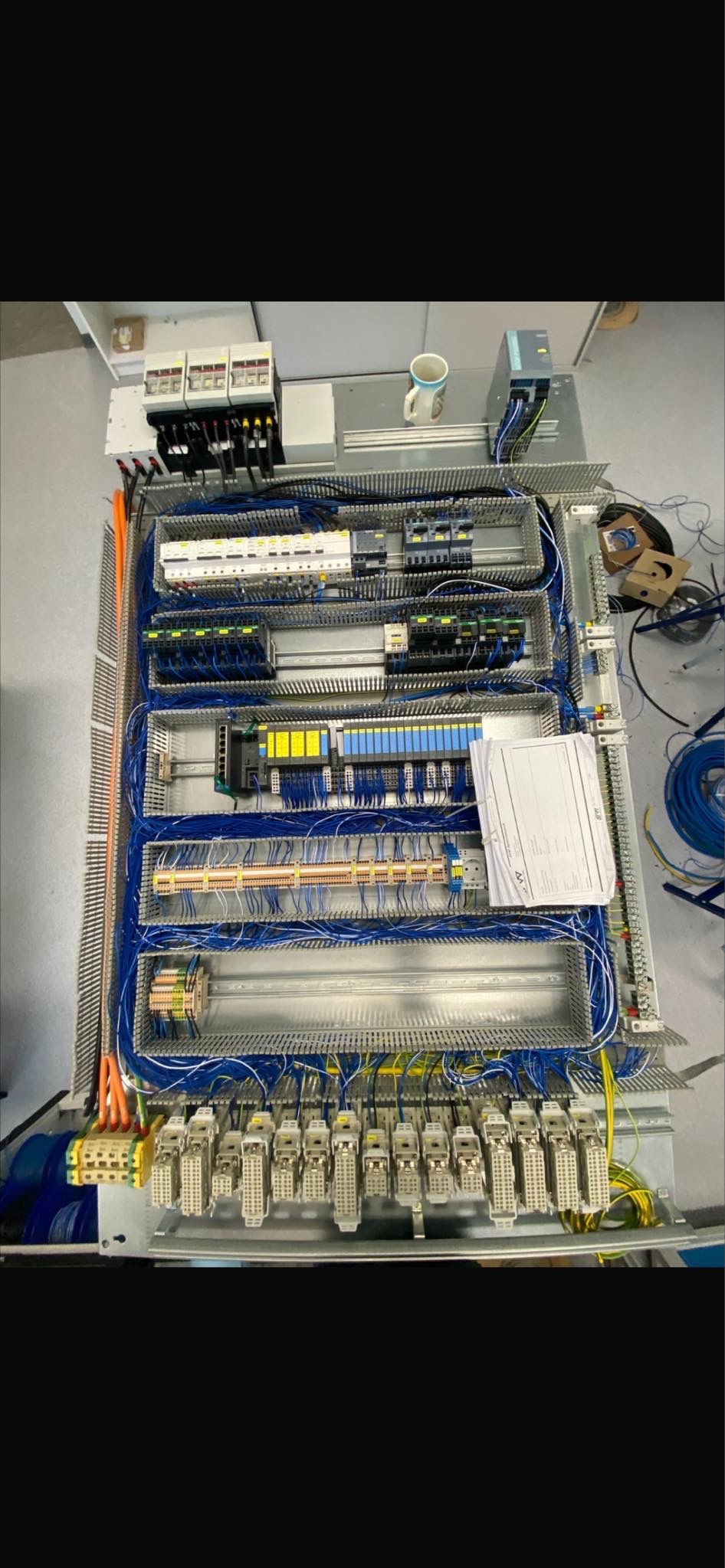 Rozbudowana szafa sterownicza z licznymi modułami, okablowaniem i elementami zabezpieczeń, z widoczną dokumentacją techniczną i kubkiem na górze.