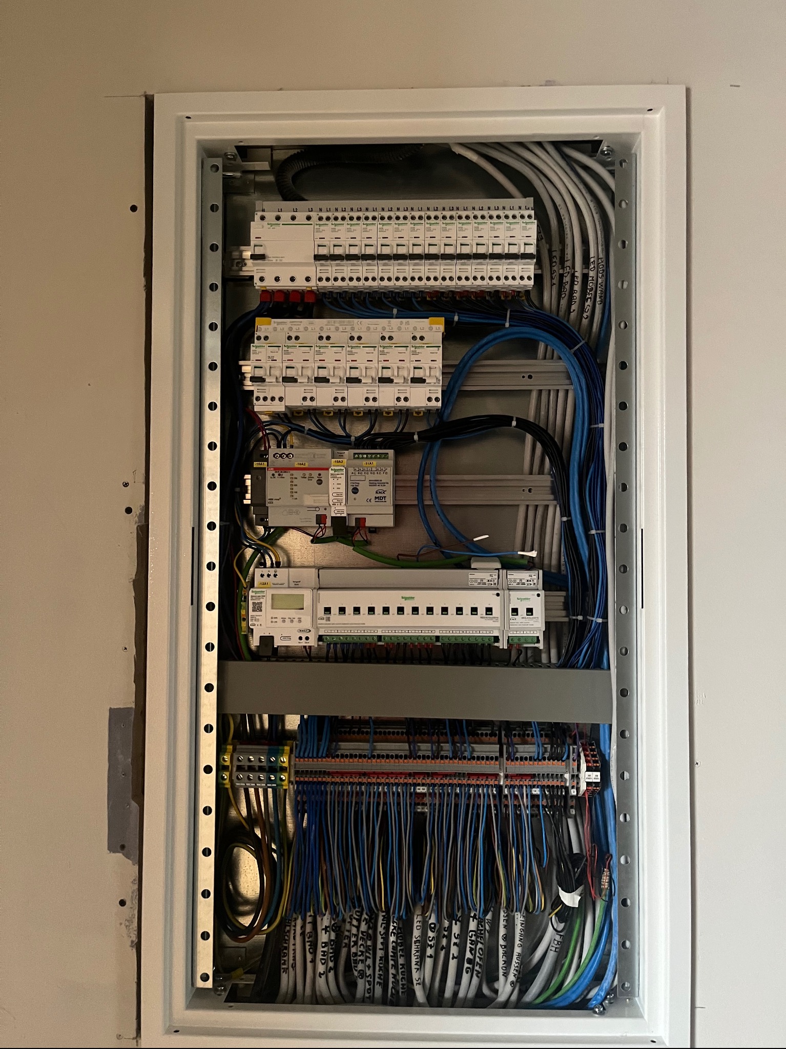 Rozdzielnia elektryczna wnękowa z zamontowanymi zabezpieczeniami, przewodami oznaczonymi opisami. Instalacja w trakcie prac, widoczne elementy montażowe.