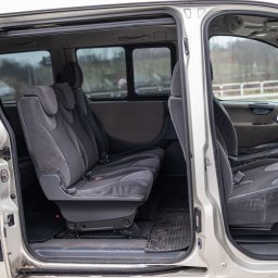 Seweryn Stodolak - Wnętrze srebrnego vana z otwartymi drzwiami, widoczne trzy rzędy szarych foteli z zagłówkami i dywaniki podłogowe.