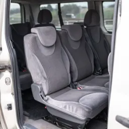 Wnętrze busa z otwartymi drzwiami, widoczne szare, welurowe fotele pasażerskie w trzech rzędach, zapięte pasy bezpieczeństwa.