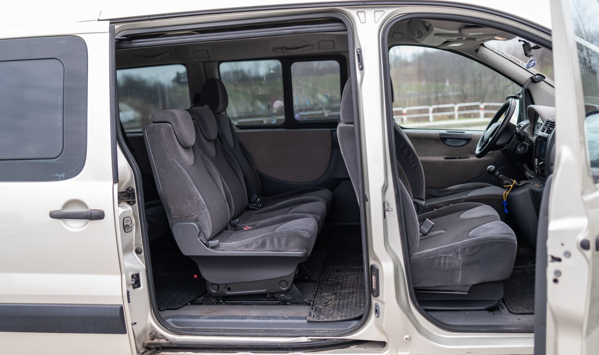 Wnętrze srebrnego vana z otwartymi drzwiami, widoczne trzy rzędy szarych foteli z zagłówkami i dywaniki podłogowe.