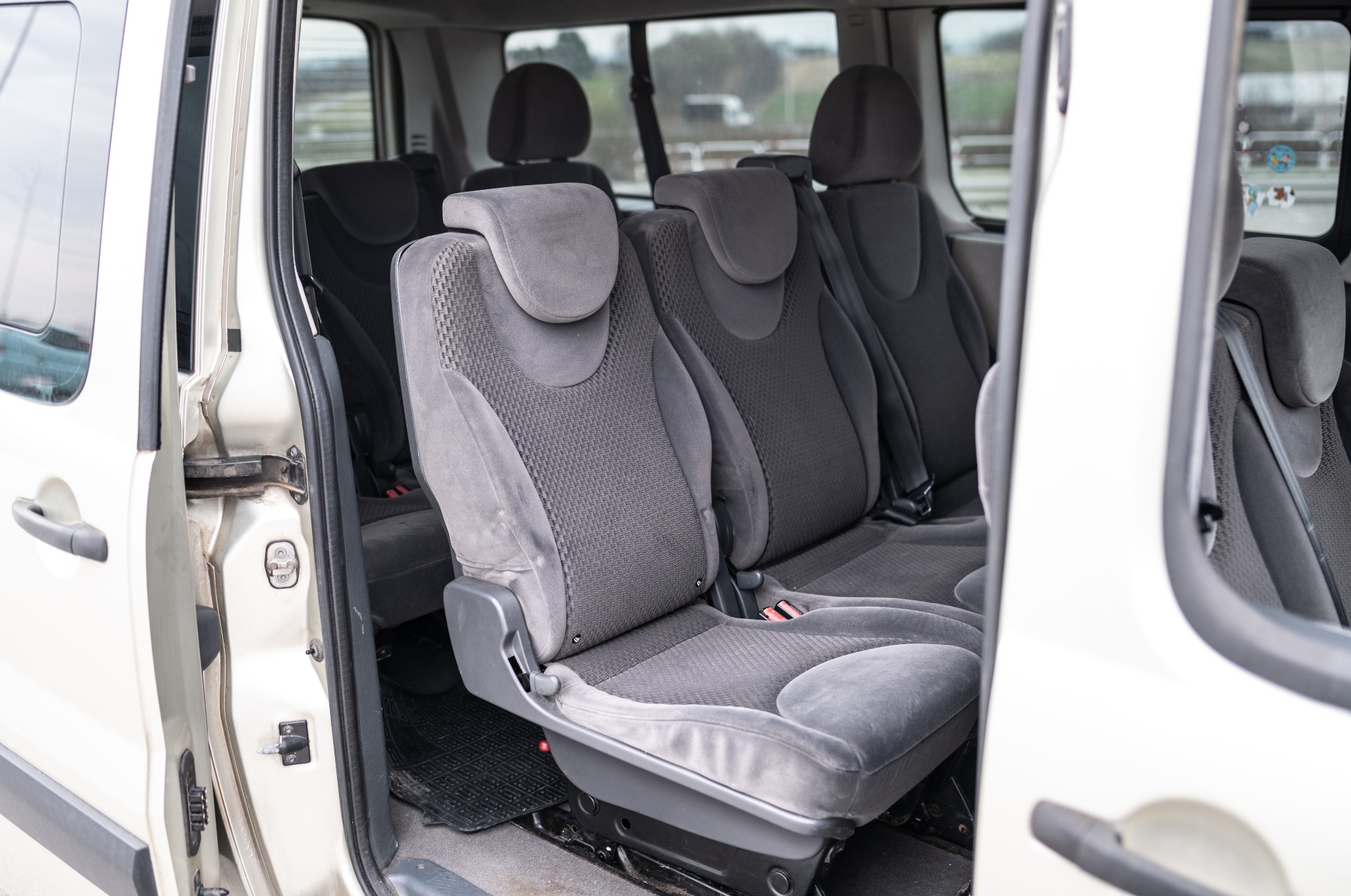 Wnętrze busa z otwartymi drzwiami, widoczne szare, welurowe fotele pasażerskie w trzech rzędach, zapięte pasy bezpieczeństwa.
