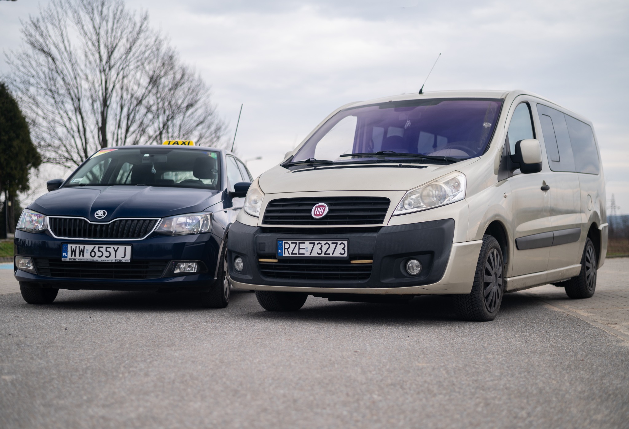 Dwa samochody: granatowa Skoda Fabia z oznaczeniem taksówki i beżowy Fiat Scudo, stoją obok siebie na parkingu w pochmurny dzień.