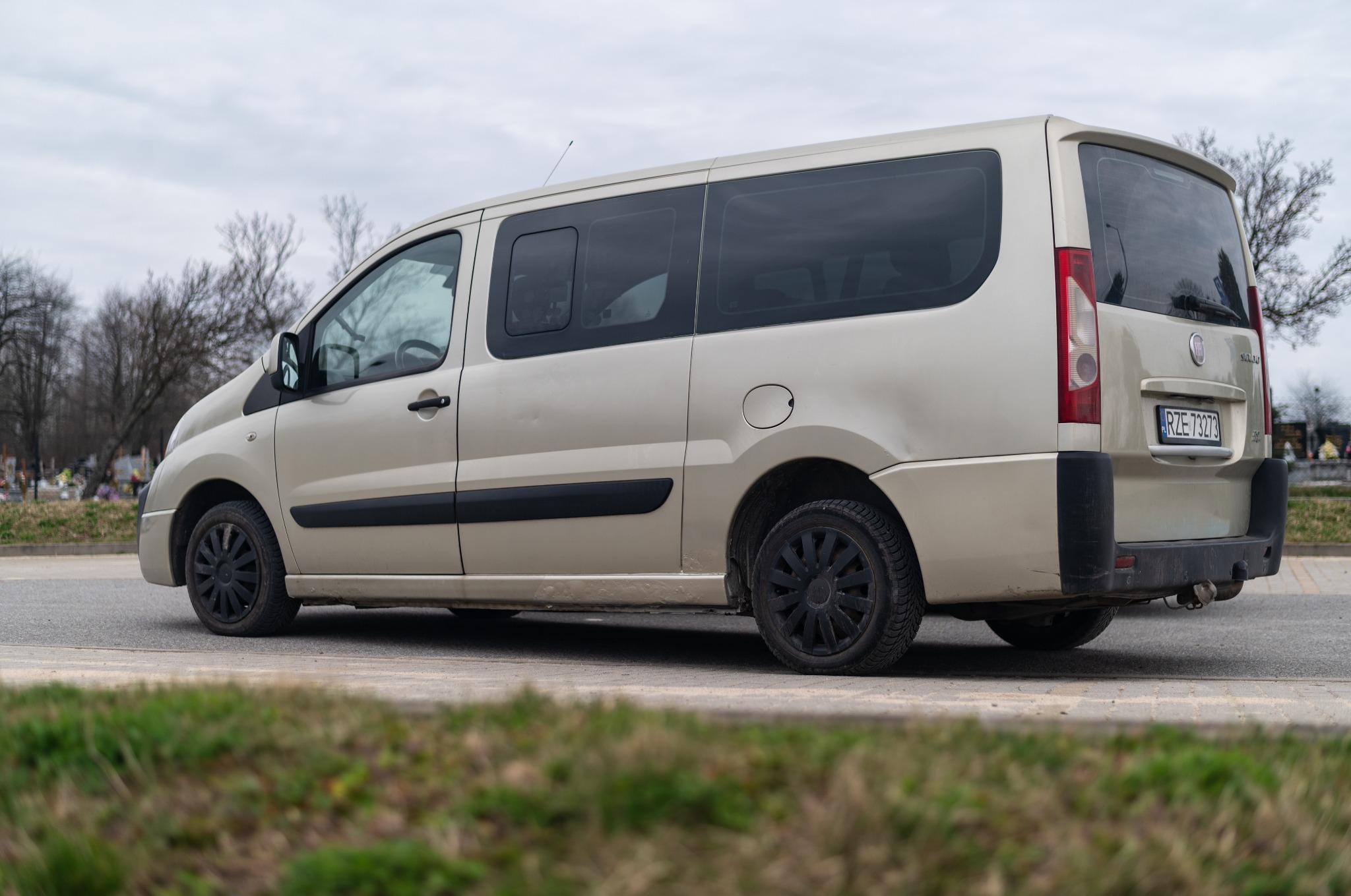 Beżowy bus na tle cmentarza, widoczny z boku i tyłu, z czarnymi felgami i tablicą rejestracyjną RZE 73273.