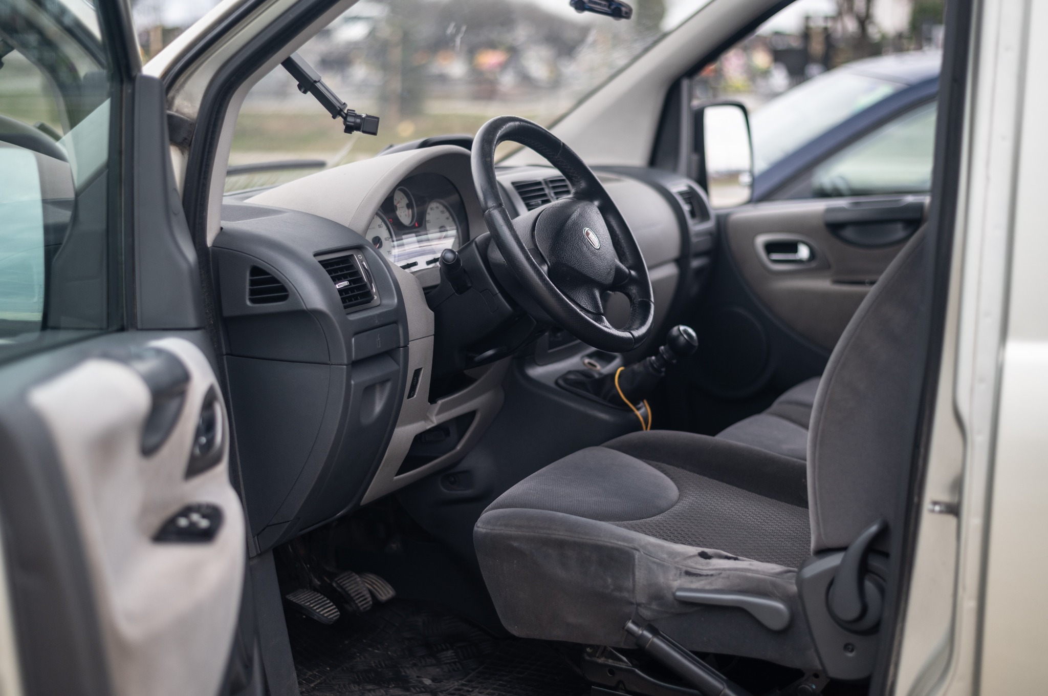 Wnętrze samochodu dostawczego z widoczną deską rozdzielczą, kierownicą i przednimi fotelami z szarej tapicerki, uchwyt na telefon na przedniej szybie.