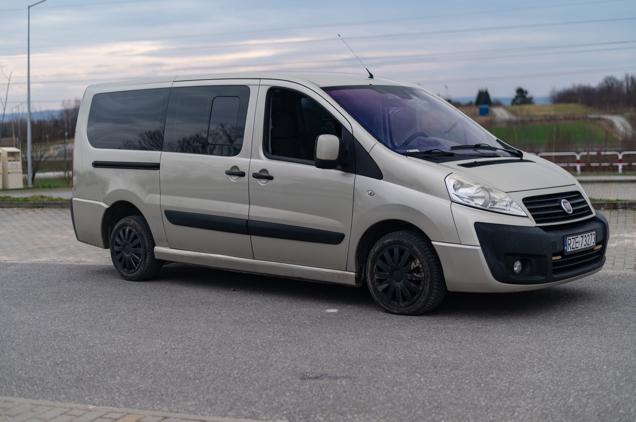 Jasny, beżowy bus Fiat Scudo z czarnymi felgami na parkingu, widok z boku, tablica rejestracyjna RZE 73273.