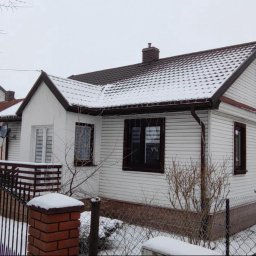 BARBAROSSA SPÓŁKA Z OGRANICZONĄ ODPOWIEDZIALNOŚCIĄ - Biały dom jednorodzinny z brązowym dachem i oknami, pokryty śniegiem, widoczny zza ogrodzenia z ceglanym słupkiem.