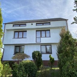 BARBAROSSA SPÓŁKA Z OGRANICZONĄ ODPOWIEDZIALNOŚCIĄ - Dwupiętrowy dom z białymi ścianami po renowacji, widoczny z poziomu ogrodu z iglakami, z brązowymi ramami okiennymi i ciemnym dachem. Widoczne niedoskonałości w strukturze tynku.