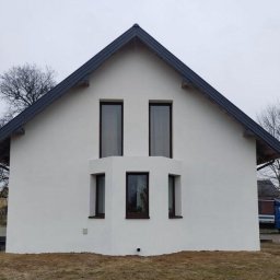 BARBAROSSA SPÓŁKA Z OGRANICZONĄ ODPOWIEDZIALNOŚCIĄ - Biały, dwukondygnacyjny dom z ciemnym dachem i ciemnymi ramami okiennymi, widziany z przodu, elewacja świeżo pomalowana, na pierwszym planie trawa. Na drugim planie widoczny samochód dostawczy.