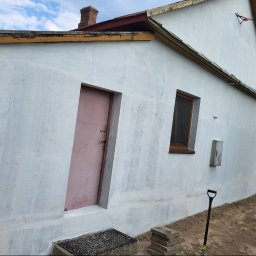 BARBAROSSA SPÓŁKA Z OGRANICZONĄ ODPOWIEDZIALNOŚCIĄ - Boczny widok na dom z otynkowaną elewacją, różowe drzwi wejściowe, małe okno z brązową ramą, widoczne ślady napraw i nierówności na ścianie, łopata oparta o ścianę, fragment dachu z ceglanym...