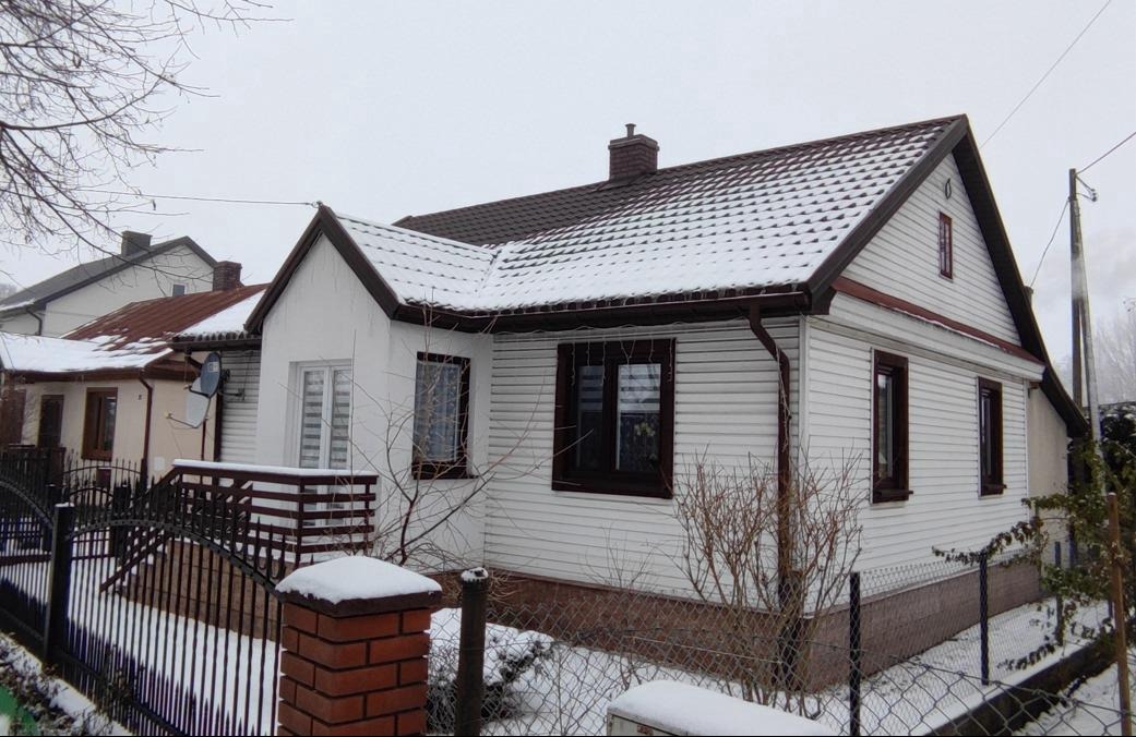 Biały dom jednorodzinny z brązowym dachem i oknami, pokryty śniegiem, widoczny zza ogrodzenia z ceglanym słupkiem.