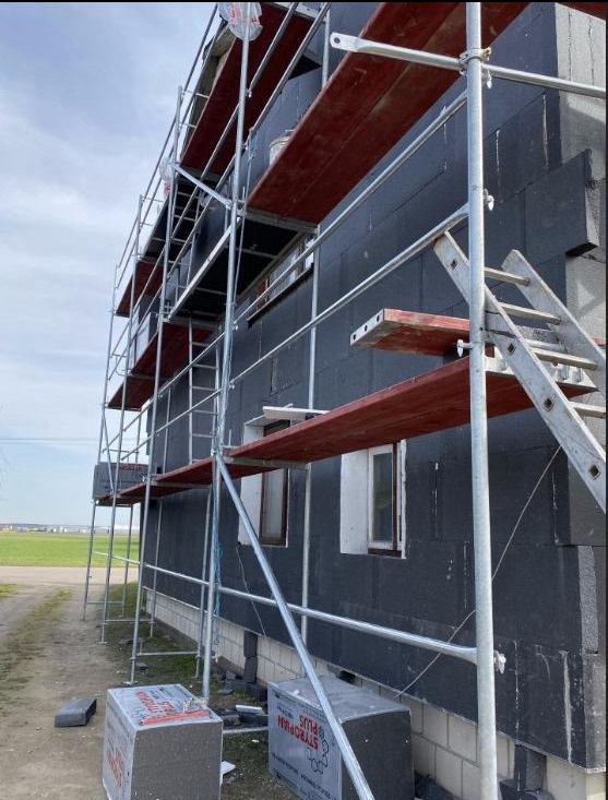 Budynek z czarną warstwą izolacyjną na ścianach zewnętrznych, otoczony rusztowaniem z drewnianymi pomostami, widoczne okna i dwie kostki styropianu na ziemi.