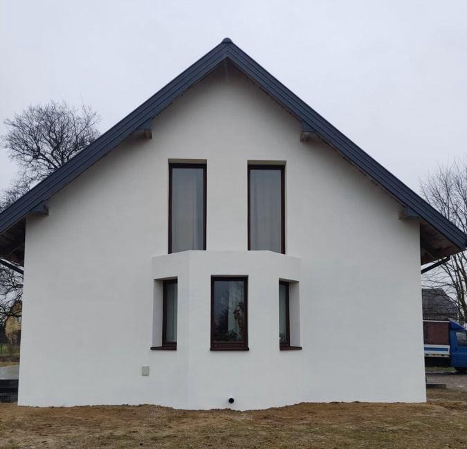 Biały, dwukondygnacyjny dom z ciemnym dachem i ciemnymi ramami okiennymi, widziany z przodu, elewacja świeżo pomalowana, na pierwszym planie trawa. Na drugim planie widoczny samochód dostawczy.