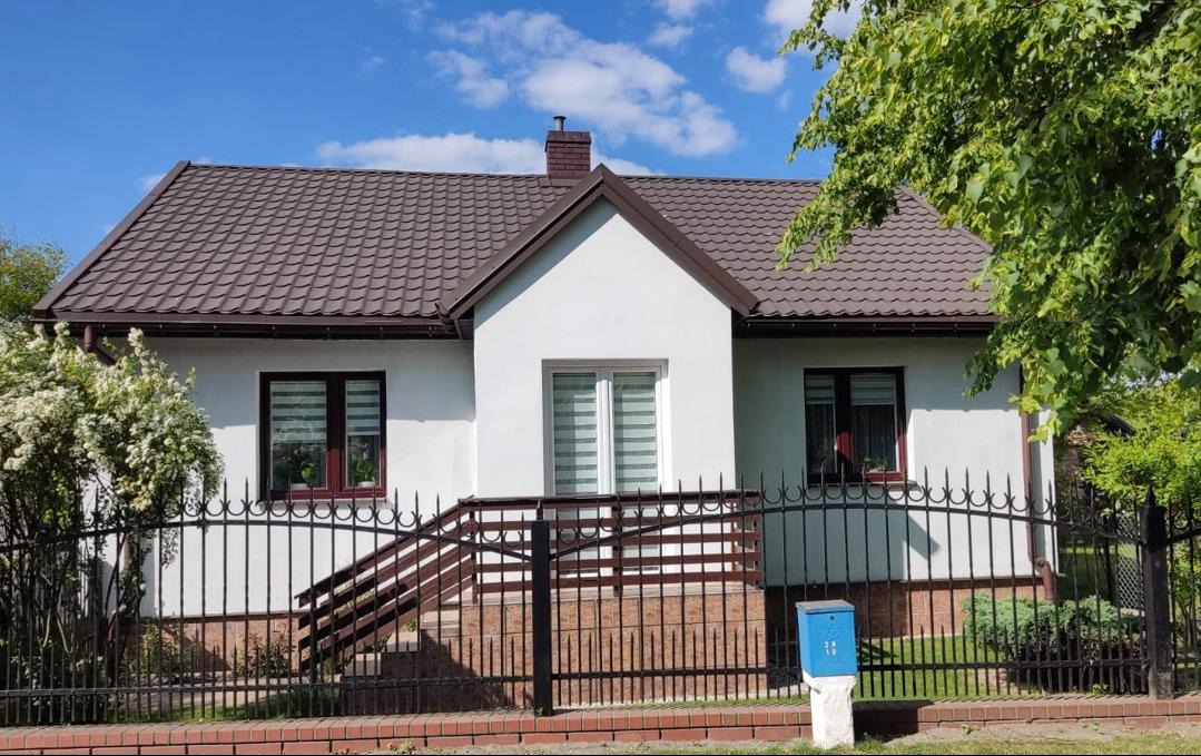 Gotowy, otynkowany dom jednorodzinny z brązowym dachem i czarnym, kutym ogrodzeniem oraz niebieską skrzynką na listy, widoczny w słoneczny dzień.