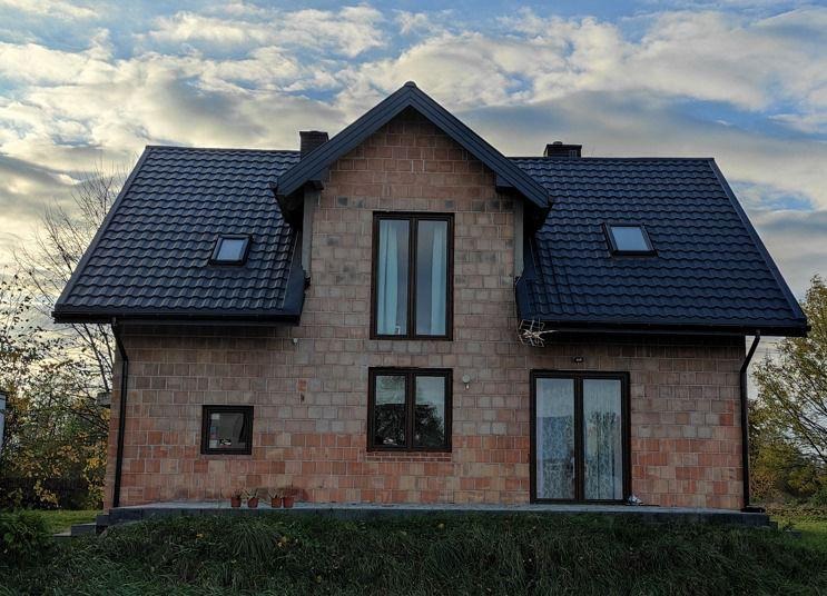 Surowy stan domu murowanego z czarną dachówką, widoczne okna w ciemnych ramach i niewielkie doniczki z kwiatami na podmurówce.