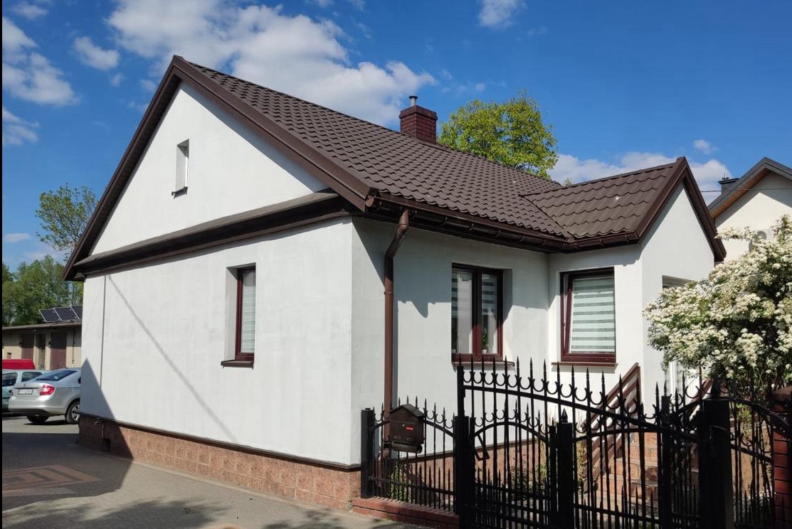 Biały dom jednorodzinny z brązowym dachem, widoczne okna z roletami, metalowe ogrodzenie i kwitnący krzew, w tle zaparkowane samochody i błękitne niebo.