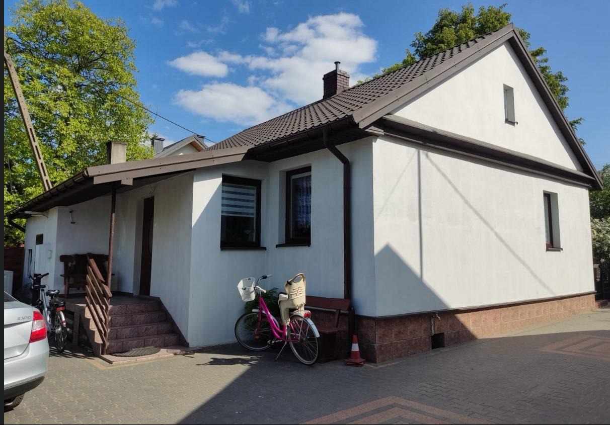 Biały dom murowany z brązowym dachem i oknami, widok z boku, z rowerem dziecięcym zaparkowanym obok ściany, na pierwszym planie kostka brukowa i fragment samochodu.