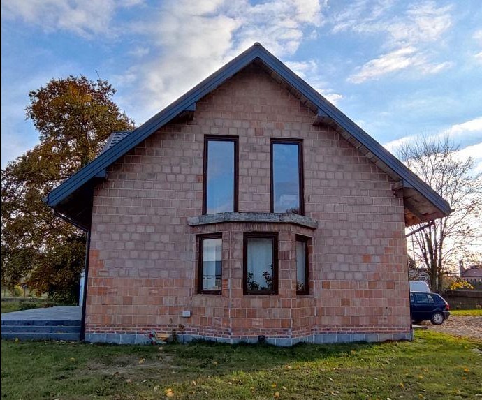 Niedokończony dom jednorodzinny z czerwonej cegły, z ciemnymi oknami i dachem, widoczny samochód w tle, jesienne drzewo po lewej stronie.
