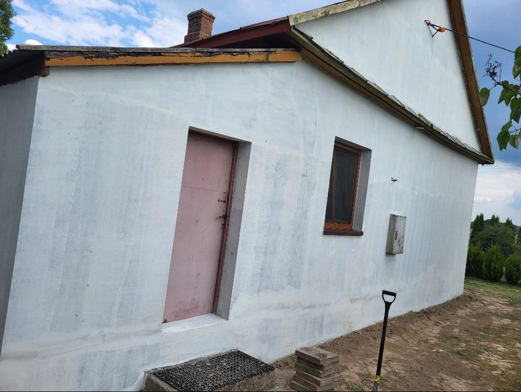 Boczny widok na dom z otynkowaną elewacją, różowe drzwi wejściowe, małe okno z brązową ramą, widoczne ślady napraw i nierówności na ścianie, łopata oparta o ścianę, fragment dachu z ceglanym...