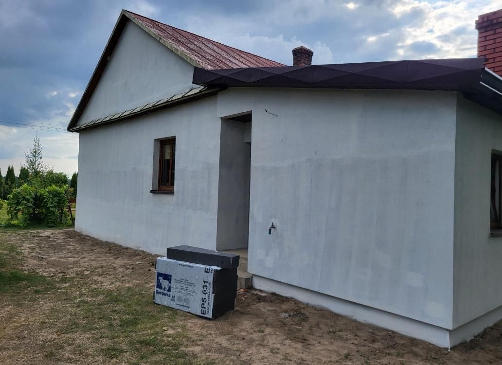 Dom jednorodzinny z otynkowaną elewacją w trakcie prac, widoczne płyty styropianowe EPS 031 na ziemi, okno z brązową ramą i czerwona dachówka.