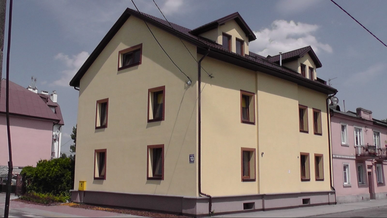 Dwupiętrowy dom murowany z beżową elewacją i brązowymi ramami okiennymi, widoczny z zewnątrz, z numerem '13' na ścianie, sąsiedni budynek w tle.