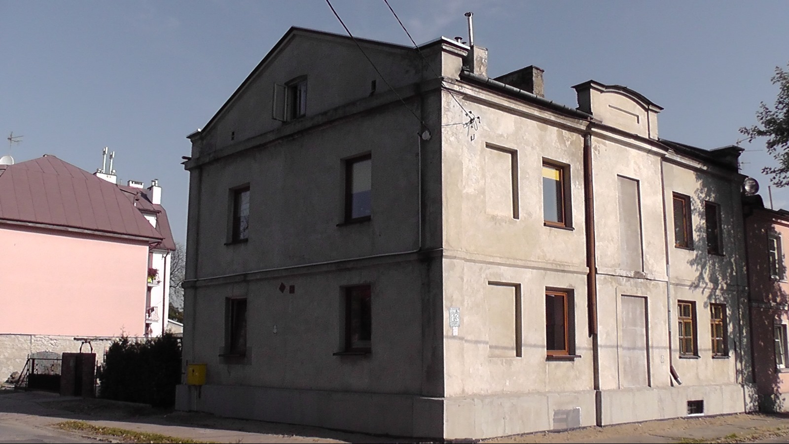 Dwupiętrowy, szary budynek mieszkalny z widocznymi śladami starzenia i brakiem ocieplenia, z fragmentem sąsiedniego budynku w tle.