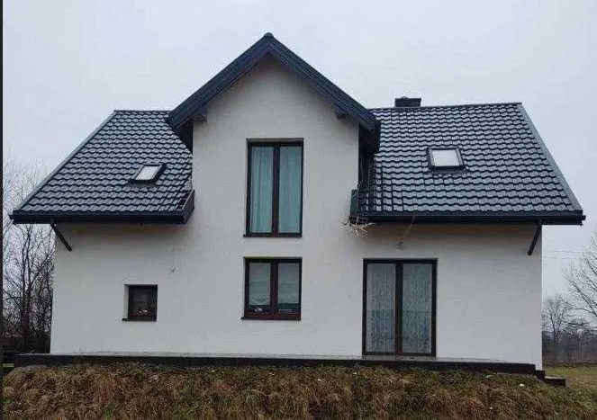 Dwukondygnacyjny dom z białymi ścianami i ciemną dachówką, widoczne okna z ciemnymi ramami i niewielkie okno dachowe, otoczenie zarośnięte trawą i krzewami, pochmurne niebo.