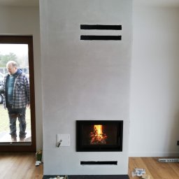 HYDRO-TECH RAFAŁ JAWORSKI - Nowoczesny kominek z paleniskiem w obudowie z jasnego betonu architektonicznego, widoczne szczeliny wentylacyjne, na podłodze torba z narzędziami, w tle osoba w drzwiach.
