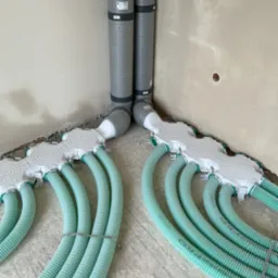 Szare rury wentylacyjne i zielone, elastyczne przewody w rozdzielaczu na surowym betonie, w rogu pomieszczenia. Instalacja rekuperacji w trakcie montażu.