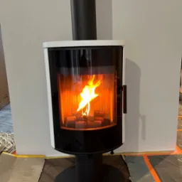 Nowoczesny kominek wolnostojący z paleniskiem, otoczony białym obramowaniem i czarnym wykończeniem, z widocznym płomieniem i przygotowanym zabezpieczeniem podłogi.