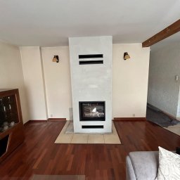 HYDRO-TECH RAFAŁ JAWORSKI - Nowoczesny kominek z czarnymi akcentami w jasnym salonie z drewnianą podłogą. Widoczne lampy ścienne i fragment sofy. Kominek w trakcie wykończenia.