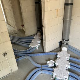 HYDRO-TECH RAFAŁ JAWORSKI - Szara rura wentylacyjna i niebieskie rury w podłodze, połączone białymi rozdzielaczami, w surowym wnętrzu budynku z białej cegły.