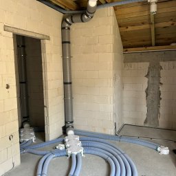 HYDRO-TECH RAFAŁ JAWORSKI - Instalacja rekuperacji w surowym budynku: szare rury pionowe i niebieskie karbowane poziome, białe rozdzielacze. Widoczne bloczki betonu komórkowego i drewniany strop.