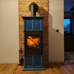 HYDRO-TECH RAFAŁ JAWORSKI - Niebieski piec kominkowy z paleniskiem, obudowany cegłą, na tle białej ściany i drewnianej podłogi. Widoczne akcesoria kominkowe.