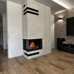HYDRO-TECH RAFAŁ JAWORSKI - Nowoczesny kominek narożny z czarnymi akcentami, płomienie widoczne przez szybę, drewniana podłoga, minimalistyczny design wnętrza z oświetleniem LED.